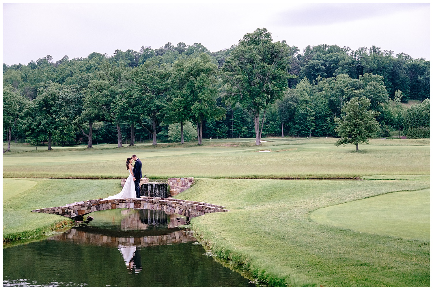 bride and groom stunning portrait on the golf course standing on a pretty stone bridge seeing their reflection in the pond